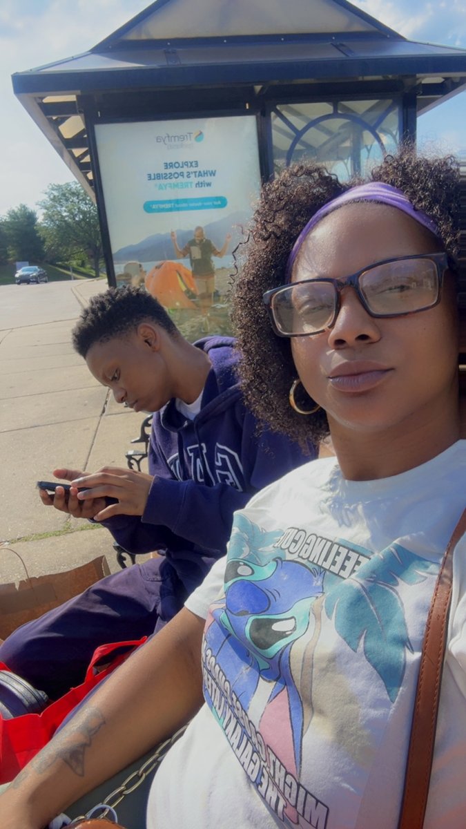 Maya on her phone at a bus stop on a sunny day